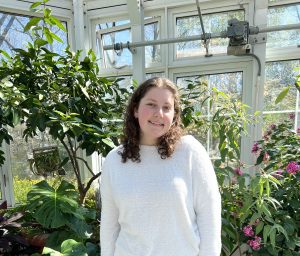 Ella Gregory wearing a white sweater and smiling in front of a greenhouse of plants