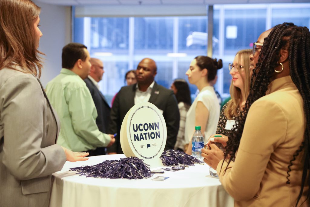 A group of UConn alumni and students gather together around a table with a UConn Nation sign, at a previous networking reception. 