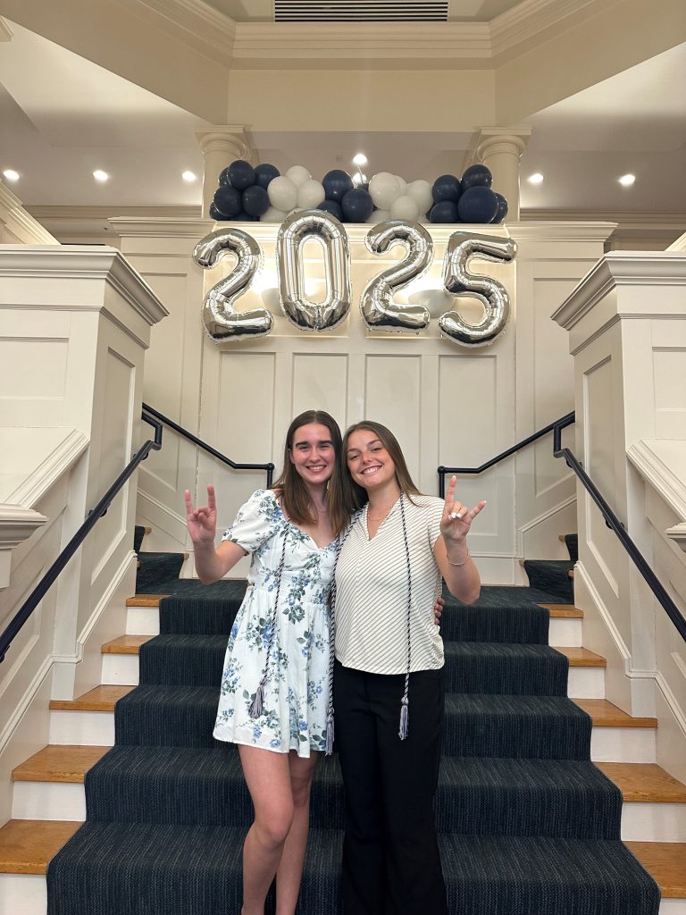 Two SAA seniors - Kaiti on left and Autumn on right - stand on the Alumni Center stairs smiling and making the husky hand signal. 2025 is spelled out behind them in balloons. 